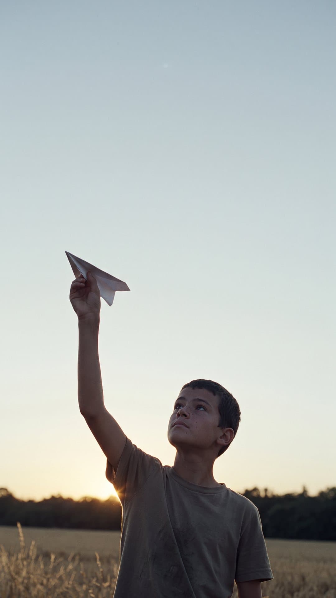 A boy in a field at dusk holds a paper plane up against the last of the light.