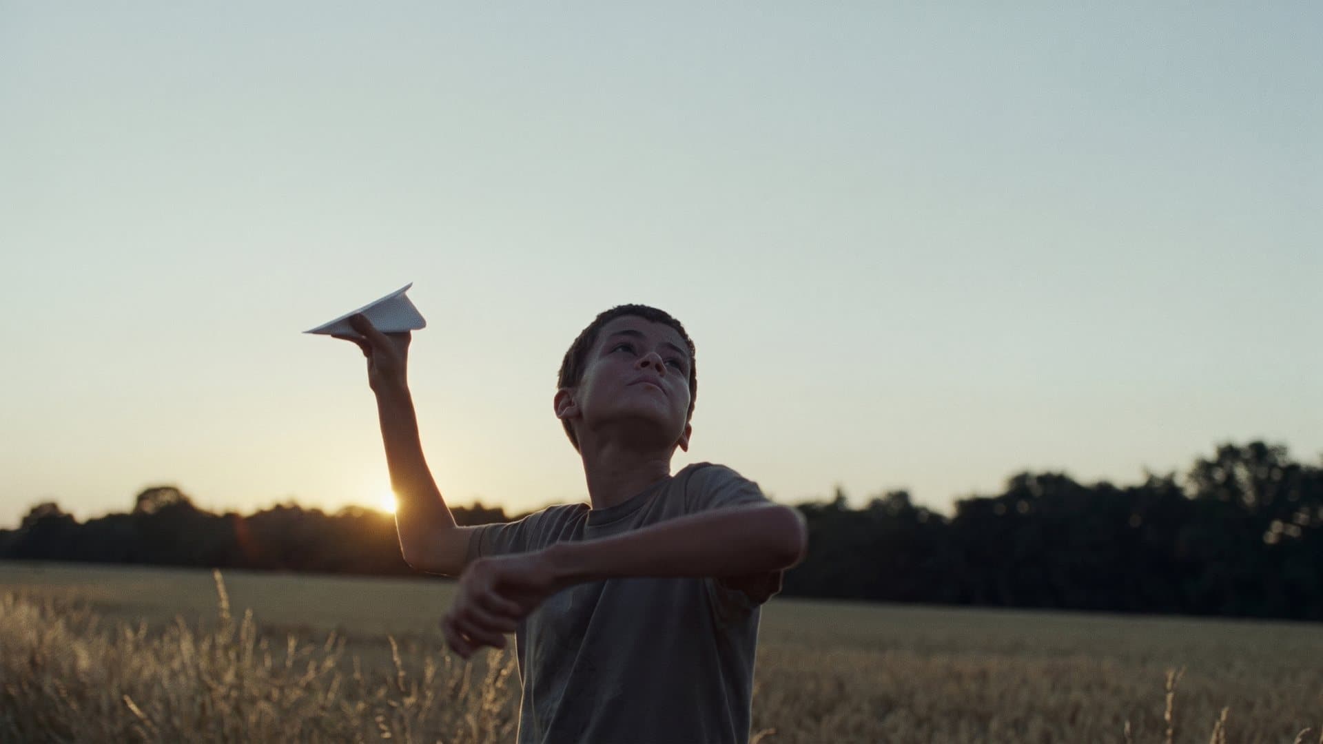 A boy in a field at dusk holds a paper plane up against the last of the light.