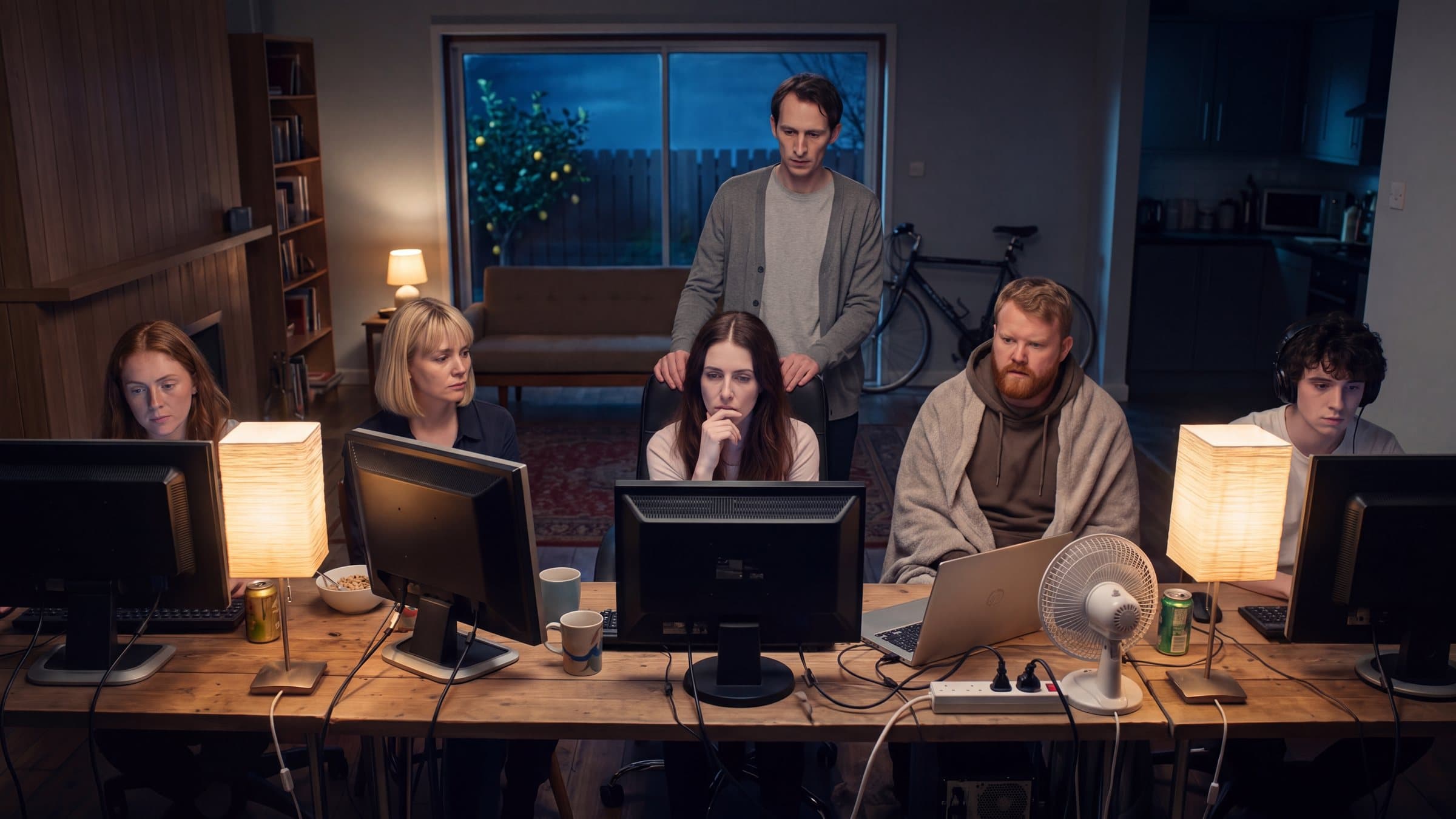 Six housemates along a long desk of monitors in a darkened living room, lit by two shaded desk lamps.